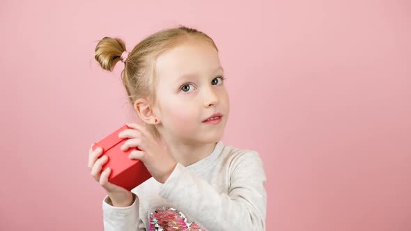 Funny Curious Little Blonde Girl Smiling and Shaking the Red Gift Box on Pink Background