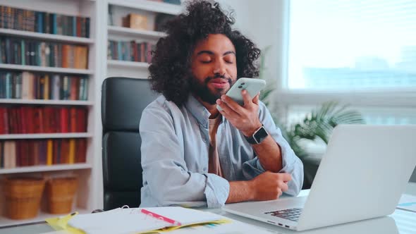 Young African American Man Smiling As Records Voice Message on Phone Sits Office