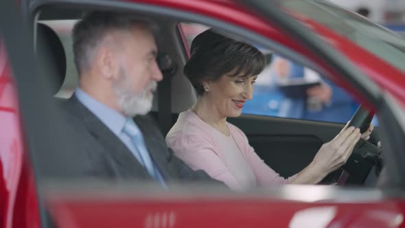 Side View of Excited Senior Beautiful Woman Sitting on Driver's Seat Talking with Blurred Man at