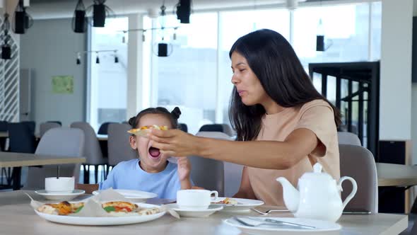 Young Mom Holds Pizza Slice and Child Girl Eats Playing