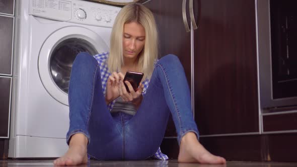 a woman is sitting on kitchen floor with her phone. Bottles with cleaning agent