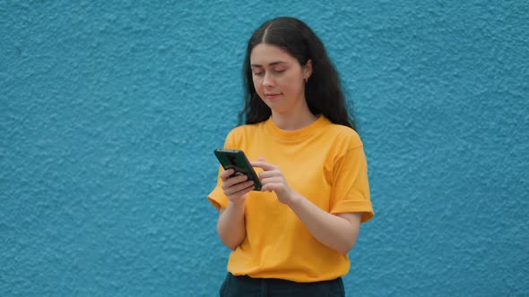 Portrait of a young caucasian woman typing on a smartphone. Blue wall on the background. Copy space.