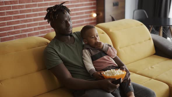 African American Father Spending Time with Small Son Sitting on Couch Eating Popcorn Watching TV