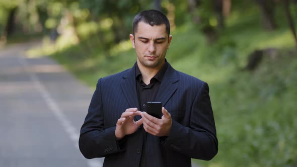 Serious Adult Caucasian Man User Stand Outdoors in Park Wears Black Formal Suit Hold Mobile