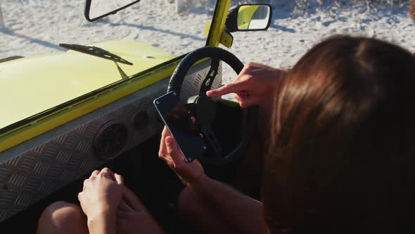 Happy caucasian couple sitting in beach buggy by the sea using smartphone