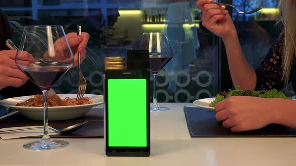 A Man and a Woman Sit at a Table and Eat, a Smartphone with a Green Screen in the Foreground