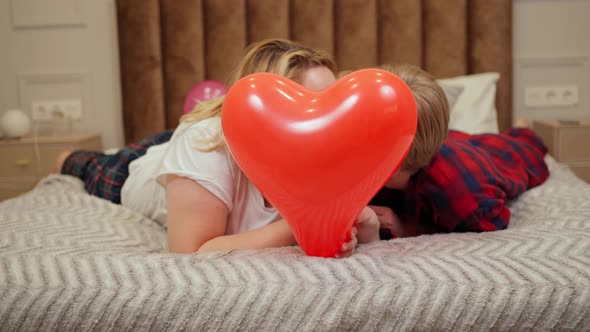 Two Women in Love Kiss and Hide Behind Heartshaped Balloons in Bed
