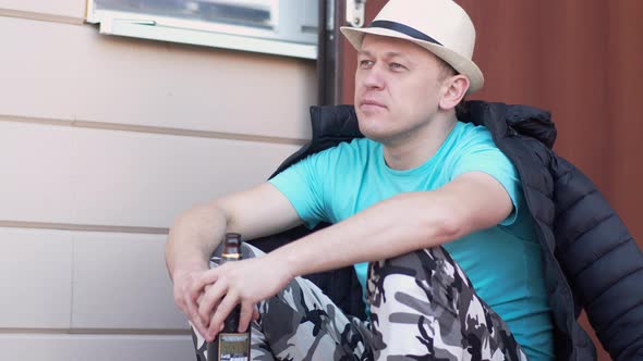 Man in a hat sits in thought with a bottle of beer on the doorstep of the house with an open door