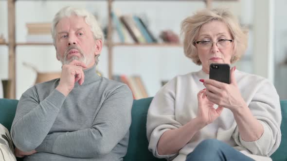 Old Woman Busy with Smartphone While Old Man is Sitting Annoyed