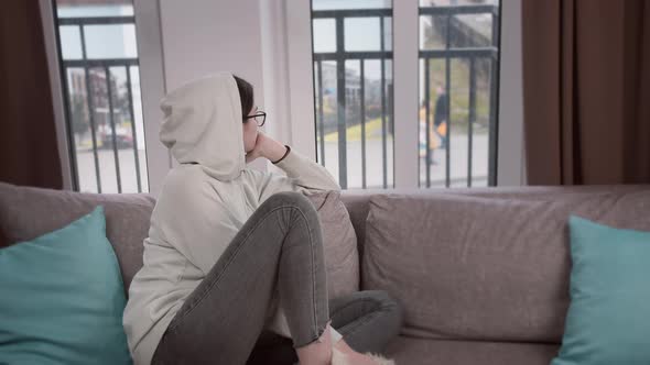 Girl in the Hood Looks Thoughtfully Out the Window While Sitting on the Sofa