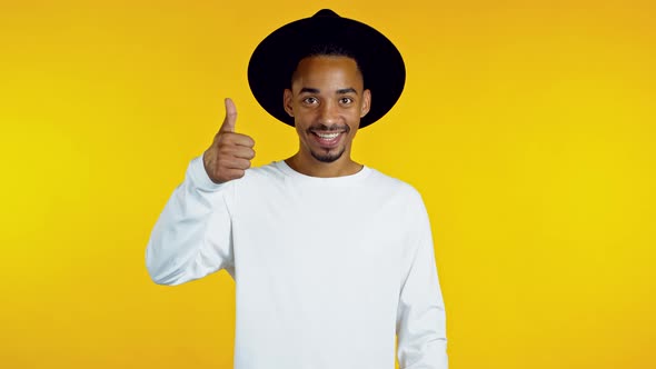 Handsome African Man in Hat Shows Thumbs Up, Like Gesture. Happy Guy on Yellow Background. Winner