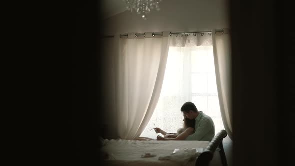 Pregnant Woman with Her Husband Sitting By the Window and Look at Ultrasound Scan of Baby on Sofa