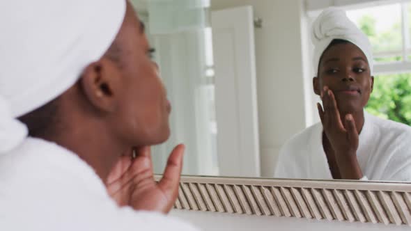 African american woman in bathrobe applying face cream while looking in the mirror at home