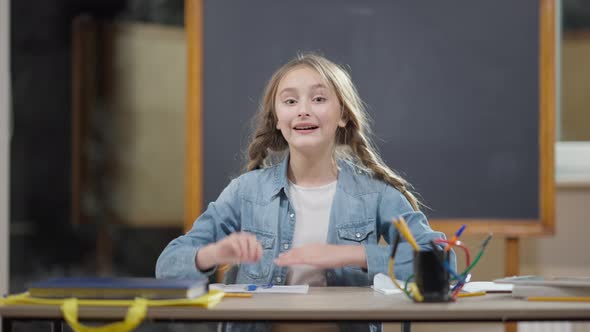 Cute Caucasian Girl with Pigtails Raising Hand Looking at Camera Sitting at Desk in Classroom