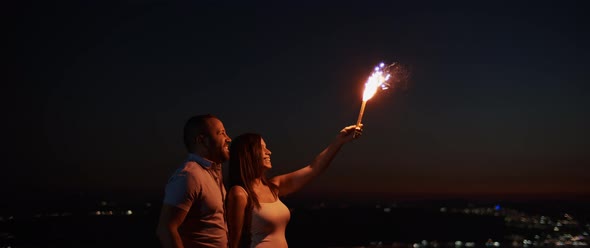 Young couple holding a firework on the hill at night, with bokeh city lights in the background