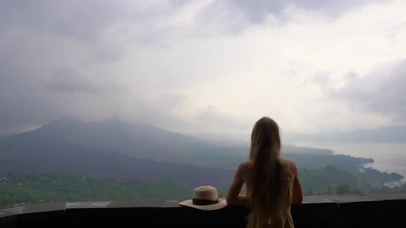 A Young Woman in a Yellow Dress and Hat Visit a Viewpoint with a View on a Mountain Batur and It's