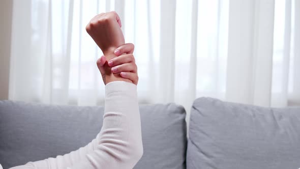 Woman is raising her hand to massage and clench her sore wrist on the sofa, using her hands to massa