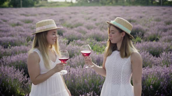 Two Caucasian Girls Twins Standing on a Lavender Field Holding Two Glasses of Red Wine Looking at