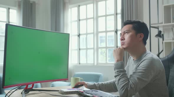 Asian Man Thinking Then Typing On Green Screen Desktop Computer While Working At Home