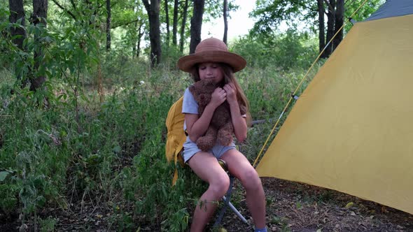 Little Girl Tourist in a Hat with Backpack and Teddy Bear Sits in the Forest Near the Tent