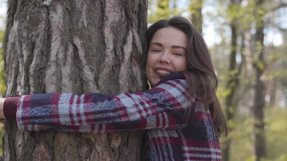 Side View Close-up of Cheerful Brunette Caucasian Girl Approaching To Tree in Forest and Hugging