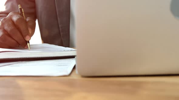 Businessman writing in dairy while using laptop