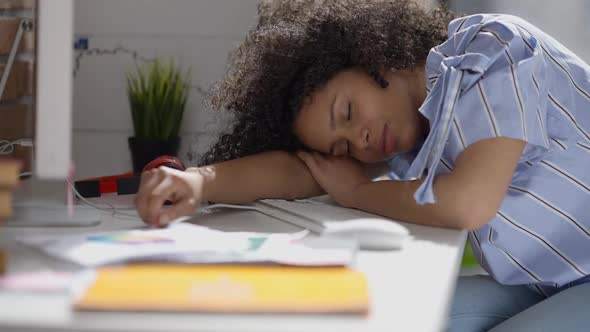Overwhelmed Young African American Woman Sleeping on Table in Home Office on Sunny Day