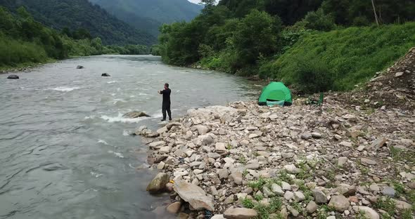 A Fisherman Fishes By Spinning a View of a Mountain River and a Green Tent