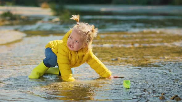 Little girl in yellow rubber boots and jacket standing in the water and plays with paper ship