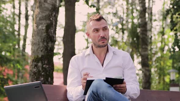 A Concentrated White Collar Man Writes Down Plans in a Notebook on a Bench