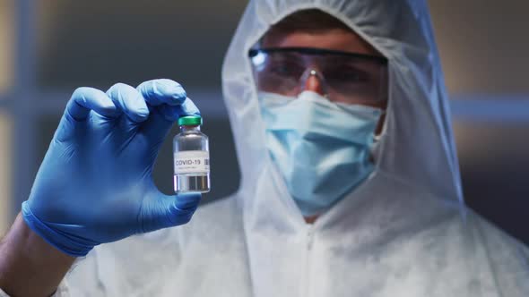 Caucasian male medical worker wearing protective clothing mask and gloves holding vaccine vial in la