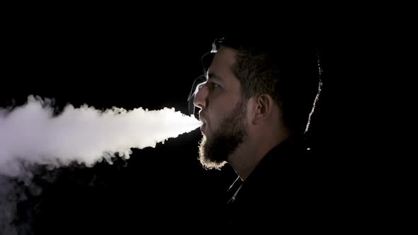 A young white bearded man in black shirt is vaping an electronic cigarette.