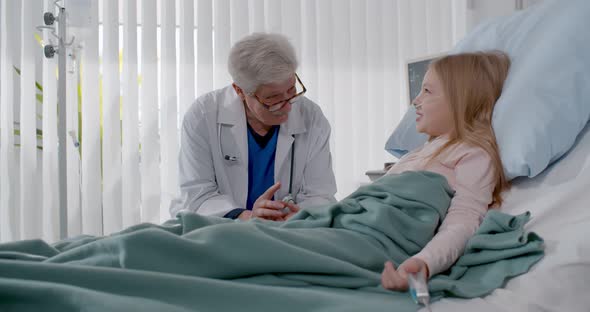 Cute Little Girl Lying in Hospital Bed and Talking with Smiling Aged Doctor