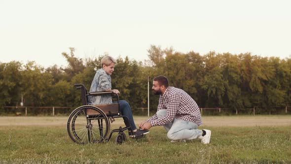 Loving man ties his shoelaces to disabled person girl sitting in wheelchair on grass in park