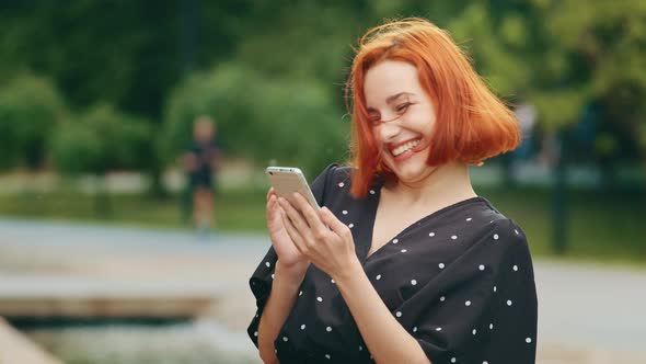 Attractive Redhead Caucasian Young Girl Woman Stands in Summer Park Looks in Mobile Phone Device