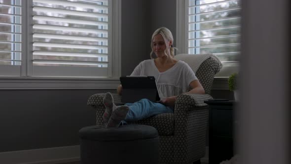 woman sitting on a chair in a corner of a reading room while scrolling on her tablet with her feet u