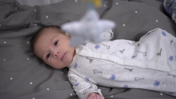 A Newborn Baby Lies in a Crib and Looks at a Toy Mobile