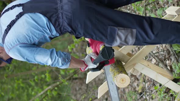 Vertical Shot Lumberjack in Workwear and Goggles Saws Tree Trunks in Forest with Electric Saw