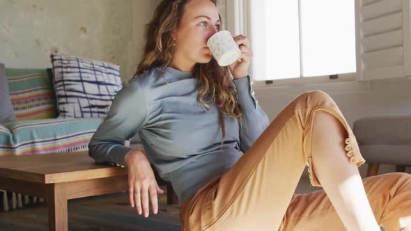 Thoughtful caucasian woman sitting on floor in living room drinking coffee, looking out of window