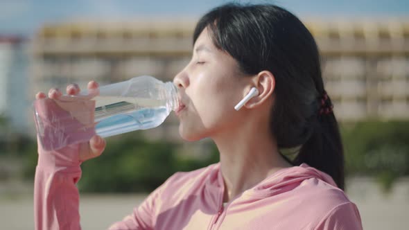Asian fitness girl drinking from sports bottle during a workout pause at a beautiful beach.
