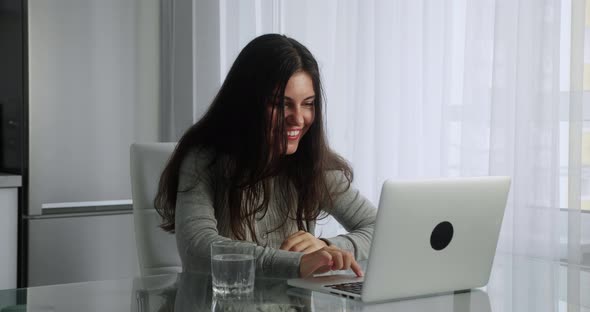 Young Brunette Woman with Long Hair Have Video Chat in Her Laptop Sitting of Kitchen Interior