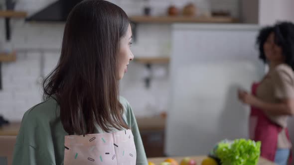 Beautiful Asian Woman in Apron with Toothy Smile Looking Back at African American Friend Writing