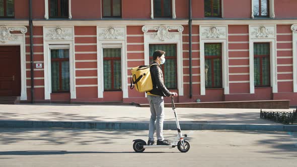 Delivery Man with Yellow Bagpack and Protective Mask Riding Electric Scooter Through the City with