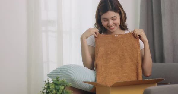 Happy Beautiful Asian Woman Opening Box With Ordered Clothes At Home On Couch.