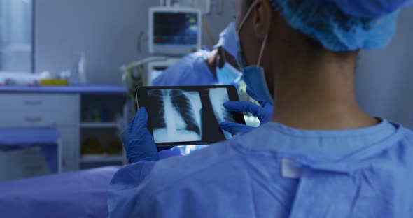 Mixed race surgeons wearing protective clothing looking at lung x-ray on tablet in operating theatre