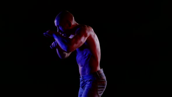 a Muscular Bald Man with a Bare Torso and Jeans Strikes a Combination of Blows on a Black Background