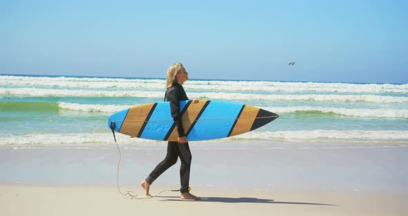 Side view of active senior Caucasian female surfer walking on beach in the sunshine 4k