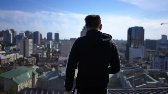 Back View Young Man Walking to Glass Fence on Rooftop Looking at Beautiful Kyiv Downtown in Ukraine