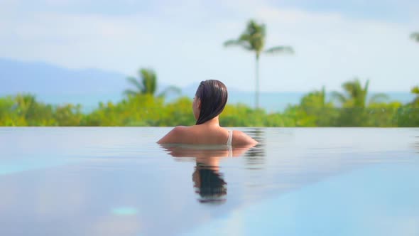 An unrecognizable woman inside infinity pool looking at amazing tropical greenery on hills backgroun