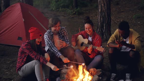 Young Lady Traveler Is Playing the Guitar, Her Friends Are Cooking Food on Fire and African American